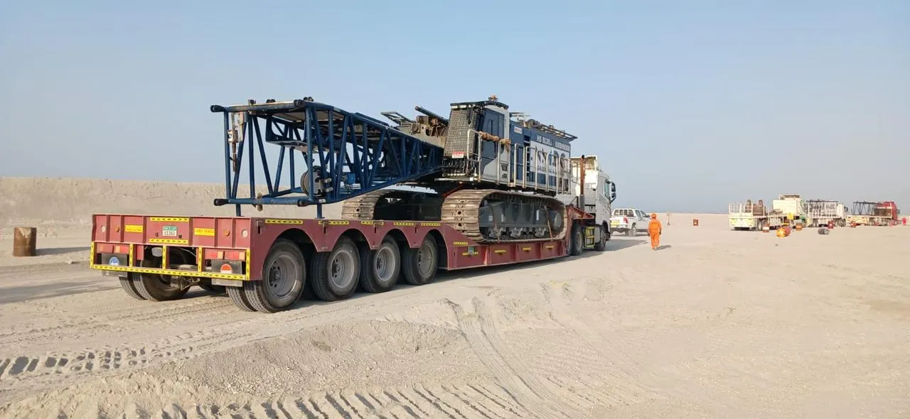 Large blue piling rig loaded on a low-bed trailer being moved across a desert site in the UAE