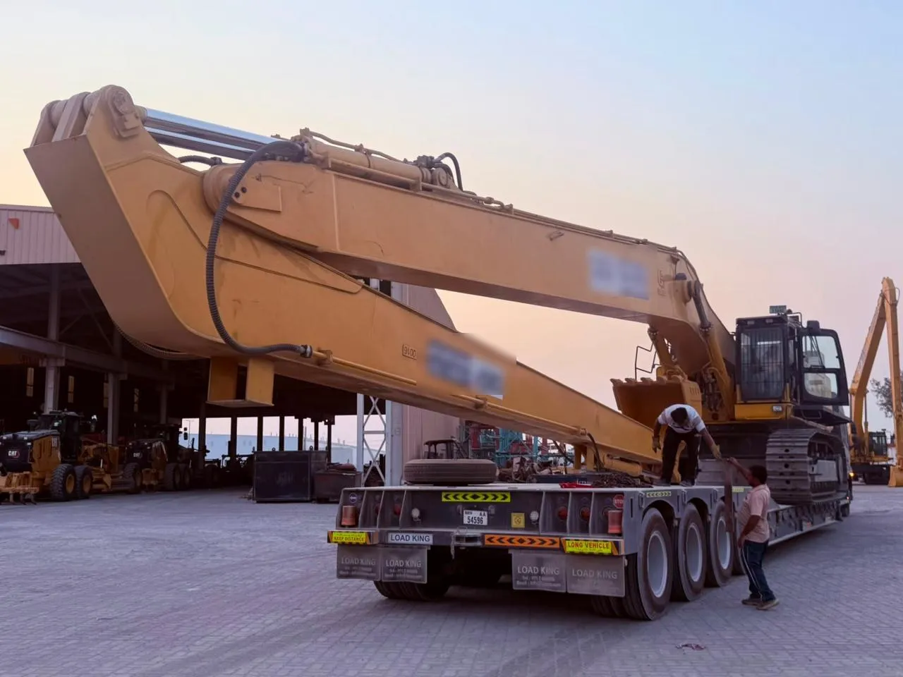 Crew fastening chains on a long-reach excavator loaded on a lowbed trailer at dusk