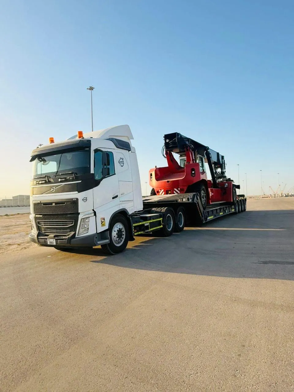 Volvo prime mover hauling a red reach stacker on a low-bed trailer at a port yard
